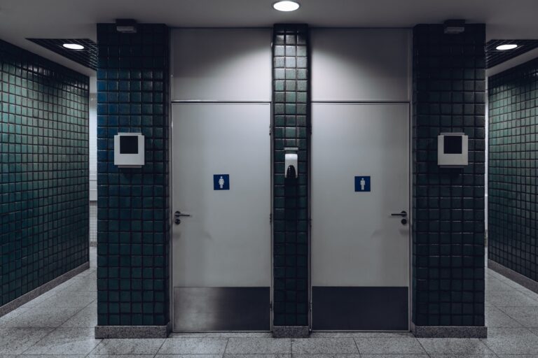 Dark public toilet entrances for men and for women in a modern airport terminal building or railway station depot, with greenish ceramic tiles on the wall: two dors and two more entrances