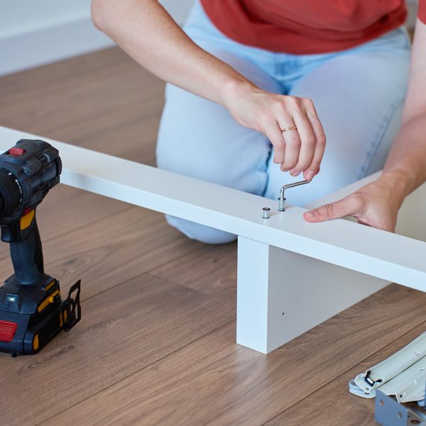 Woman assembling bed frame on wooden floor, using wrench and drill for installation. DIY furniture assembly and home improvement